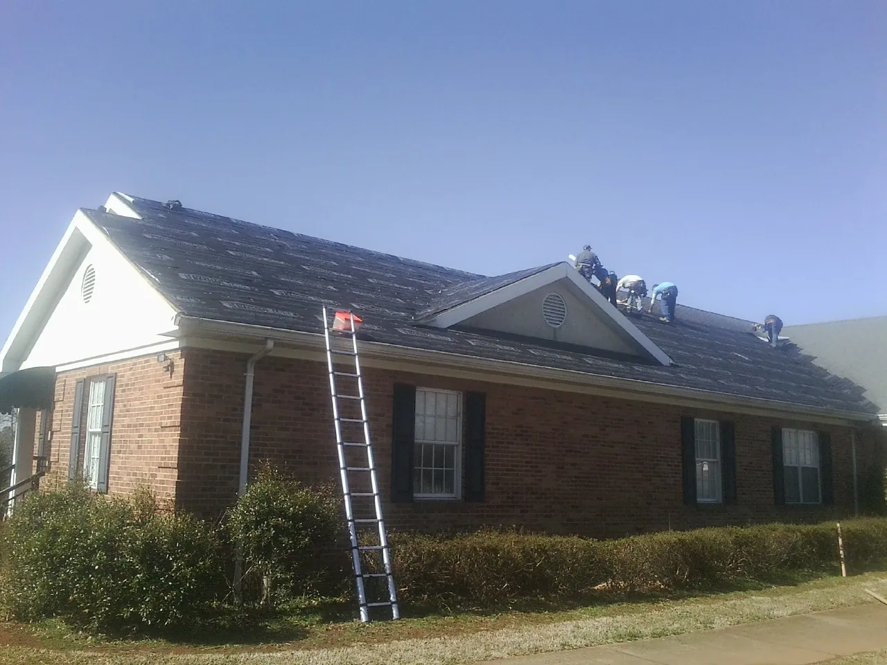 Crew tearing shingles off a brick ranch roof with a ladder set — Metal Roof Repair work by our crew in Salina